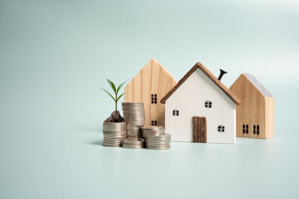 Three mini wooden homes standing next to each other with a multiple stacks of coins placed in front of them