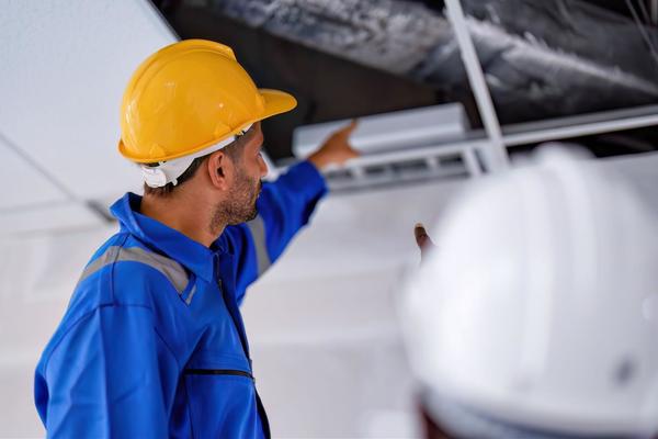 An HVAC tech wearing a yellow hard hat and blue jacket is pointing to ductwork in a ceiling